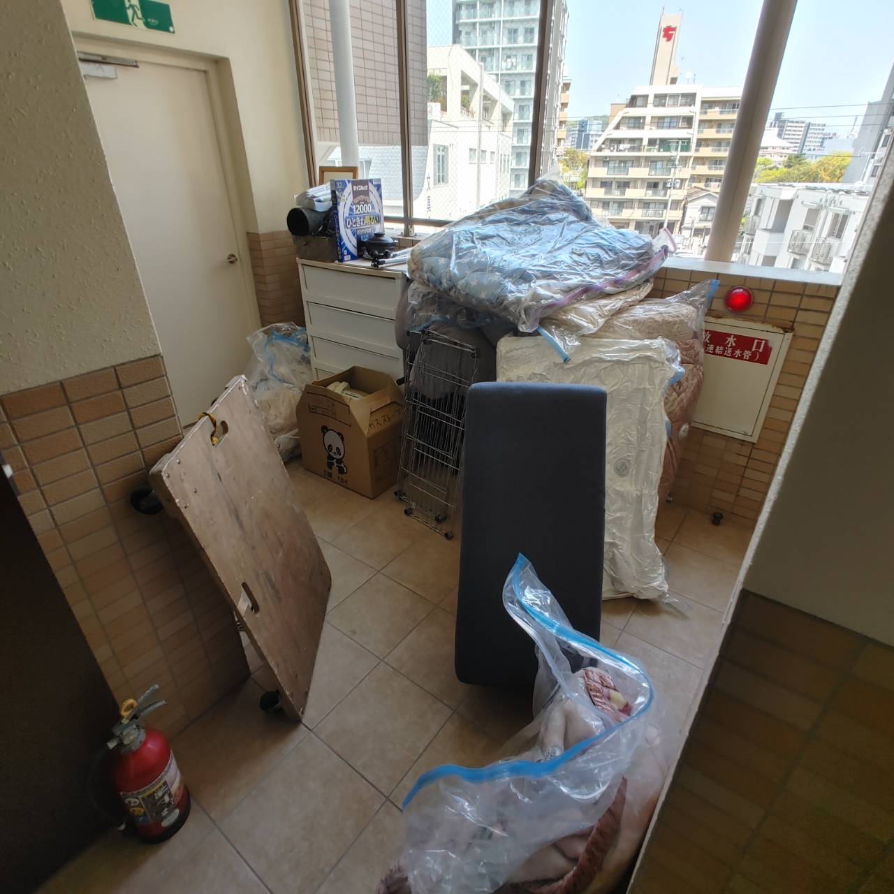 鹿児島市上荒田町のマンションベランダに放置された不用品（布団、ベッド、マットレス、チェスト、健康椅子、自転車など）の回収前の様子。鹿児島市の許可業者HAKKOが丸ごとお片付けします。